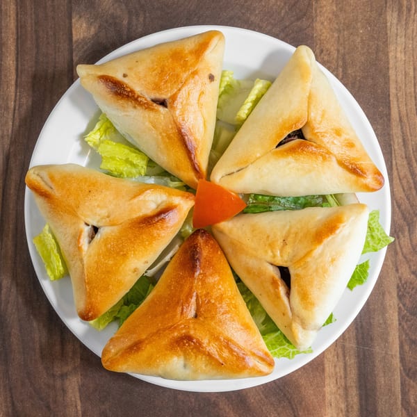 Five golden-brown triangular pastries, likely fatayer, arranged on a white plate with lettuce and a tomato wedge.