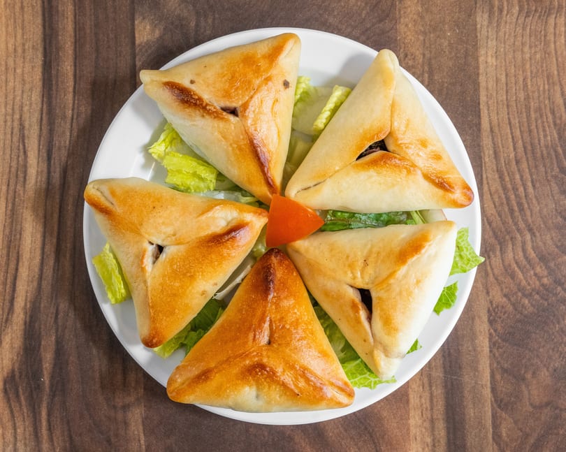 Five golden-brown triangular pastries, likely fatayer, arranged on a white plate with lettuce and a tomato wedge.