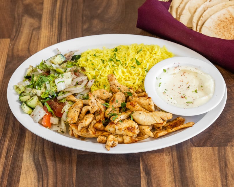 Chicken shawarma plate with yellow rice, salad, and garlic sauce, with pita bread in the background.