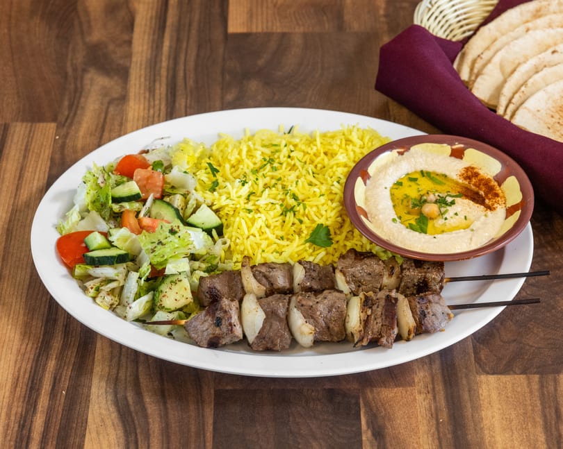 Beef shish kabob plate with yellow rice, salad, hummus, and pita bread on a wooden table.