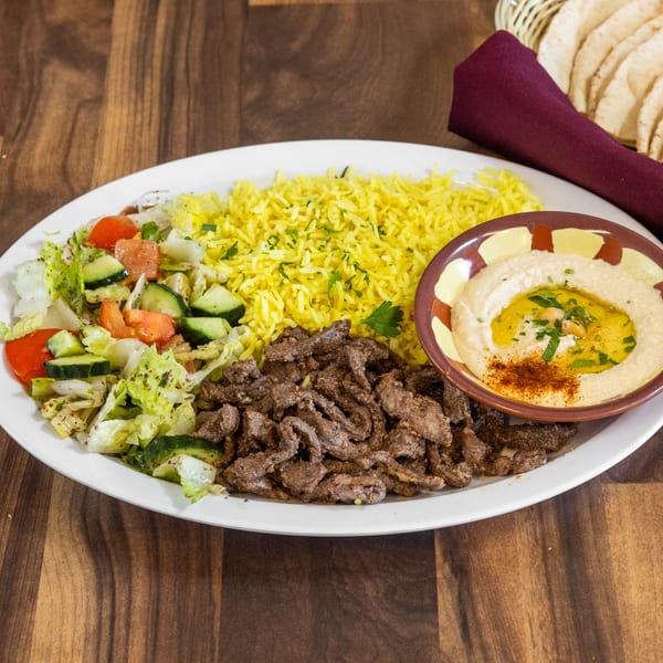 A white plate with beef shawarma, yellow rice, salad, hummus, and pita bread on a wooden table.