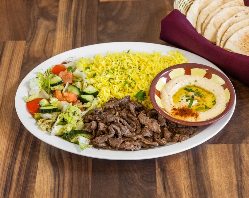 A white plate with beef shawarma, yellow rice, salad, hummus, and pita bread on a wooden table.