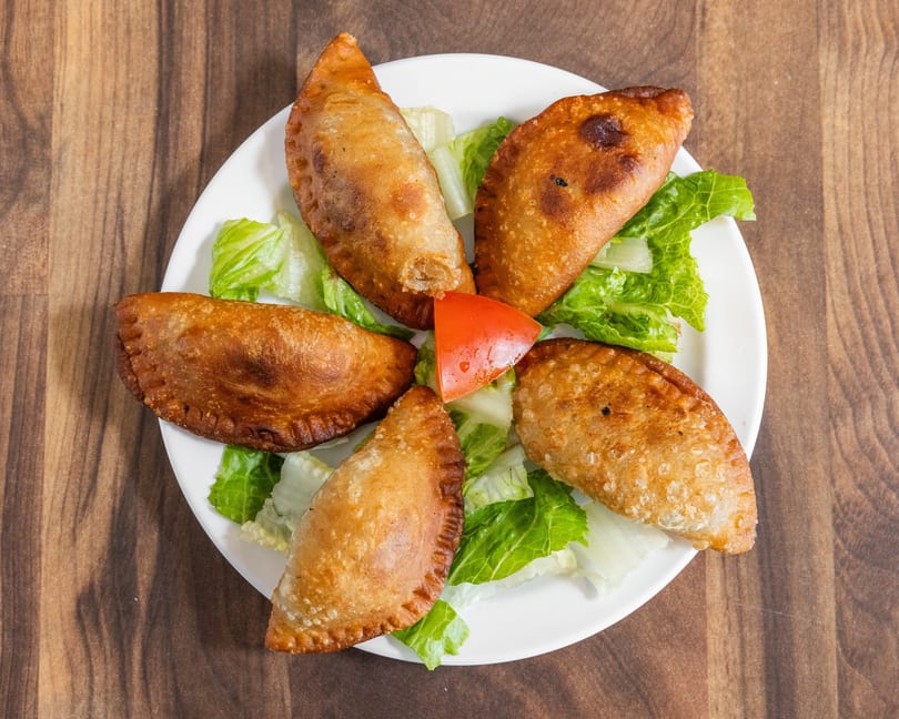 Five golden-brown empanadas arranged on a white plate with lettuce and a tomato wedge.